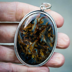 May include: Oval pendant with a polished pietersite gemstone, showcasing swirling patterns of blue, gold, and brown. The gemstone is set in a silver-colored metal frame with a loop for a chain.