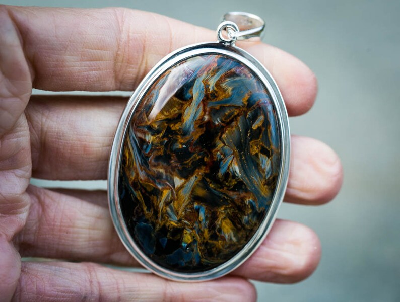 May include: Oval pendant with a polished pietersite gemstone, showcasing swirling patterns of blue, gold, and brown. The gemstone is set in a silver-colored metal frame with a loop for a chain.