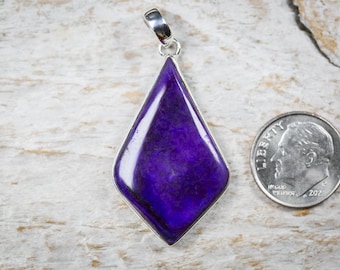 May include: A diamond-shaped, deep purple pendant with a silver bail. The pendant is next to a US dime coin for size comparison. The pendant has a polished, glossy finish and is set in a silver bezel.