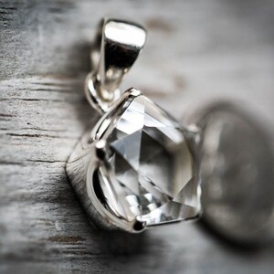 May include: A silver pendant featuring a clear, faceted, pentagonal-shaped gemstone held by a four-prong setting. The pendant includes a polished silver bail at the top for attachment to a chain, showcasing a bright, reflective metallic finish.
