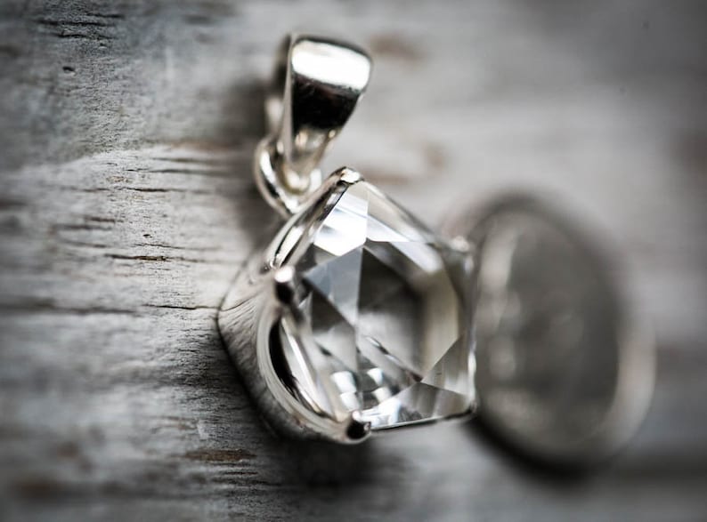 May include: A silver pendant featuring a clear, faceted, pentagonal-shaped gemstone held by a four-prong setting. The pendant includes a polished silver bail at the top for attachment to a chain, showcasing a bright, reflective metallic finish.