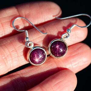 May include: A pair of silver dangle earrings. Each earring features a round, deep purple gemstone with a star-like pattern. The gemstones are set in a silver bezel and hang from a simple silver hook.