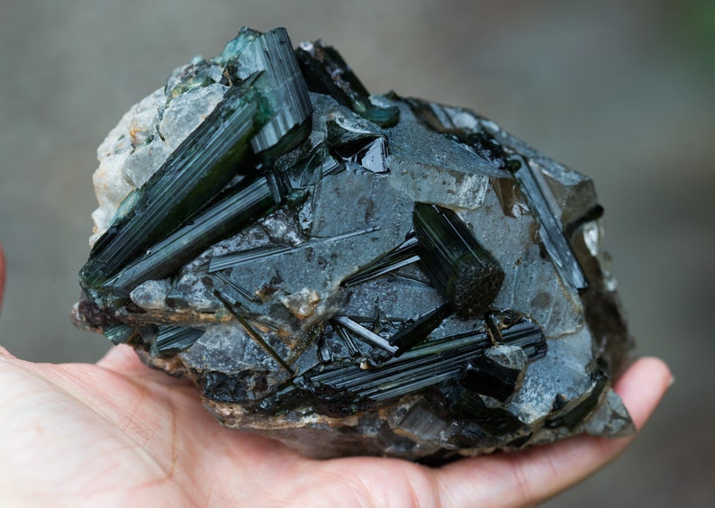 May include: A natural mineral specimen with dark green tourmaline crystals set in a grey and white stone matrix. The crystals are elongated and glassy, with a deep, rich colour. The specimen is uncut and raw.