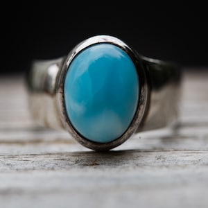 May include: A silver ring featuring a large, oval-shaped, light blue gemstone. The gemstone is set in a simple, polished silver band. The ring is displayed on a light-colored, textured surface, with a dark background.