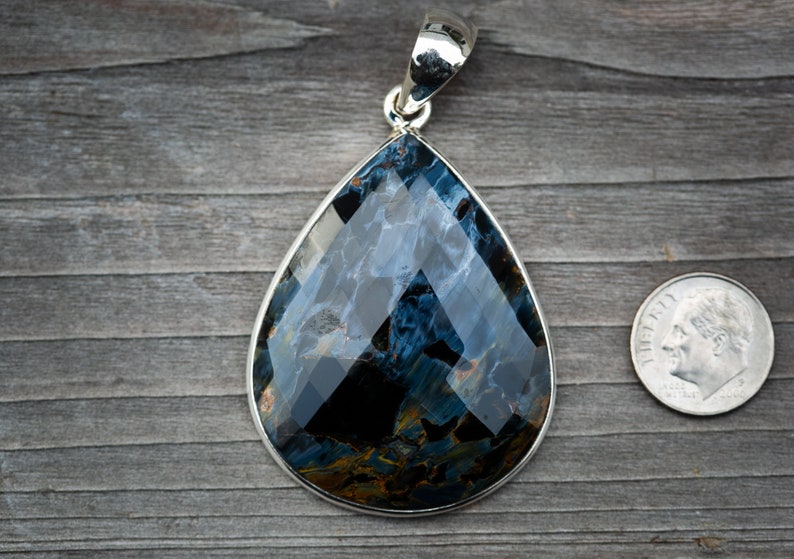 May include: A teardrop-shaped pendant with a blue and gold tiger's eye gemstone set in sterling silver. The pendant is approximately the size of a US dime.
