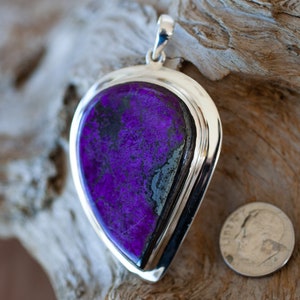 May include: A silver pendant with a large, teardrop-shaped purple and black stone. The stone is set in a silver bezel and has a polished finish. The pendant is approximately the size of a US dime.