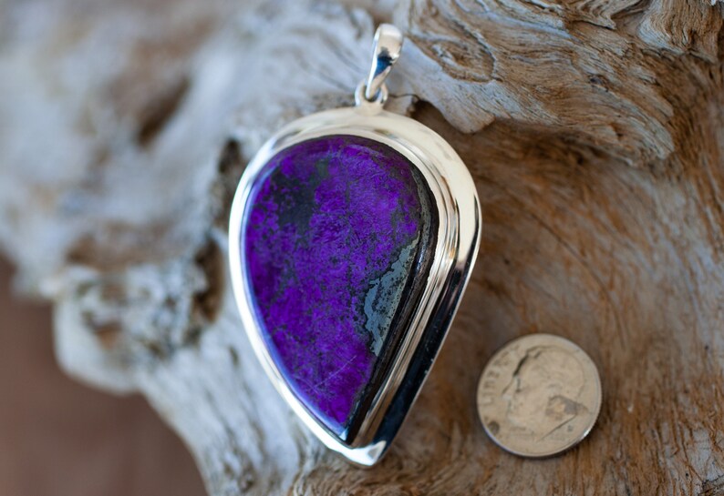 May include: A silver pendant with a large, teardrop-shaped purple and black stone. The stone is set in a silver bezel and has a polished finish. The pendant is approximately the size of a US dime.