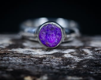 May include: A close-up of a silver ring with a large, oval-shaped, vibrant purple gemstone. The ring is set against a textured, dark wooden surface, with a blurred black background. The gemstone's color is a deep, rich purple.