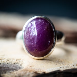 May include: A silver ring featuring a large, oval-shaped, deep purple gemstone. The polished stone is set in a simple, smooth silver band. The ring is displayed on a textured, light brown surface, with a blurred background.