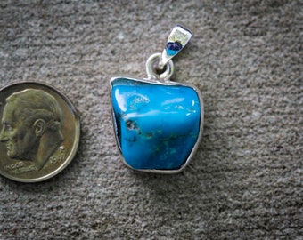 May include: A turquoise pendant with a silver setting. The pendant is a vibrant blue-green colour and has a unique, irregular shape. A U.S. dime is next to the pendant for size comparison. The pendant is on a textured grey surface.