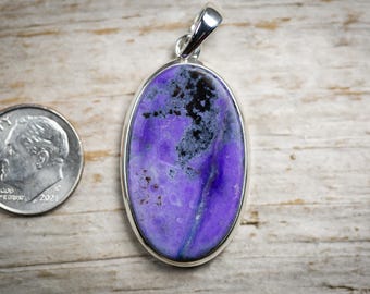 May include: Oval-shaped pendant with a vibrant purple stone, accented with black and gray patterns, set in a silver bezel. A U.S. dime is next to the pendant for size comparison. The pendant has a silver bail for a necklace.