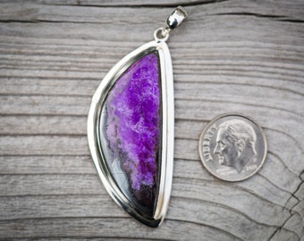 May include: A silver pendant with a large, half-circle shaped purple sugilite gemstone. The pendant is on a wooden surface next to a US dime for scale.