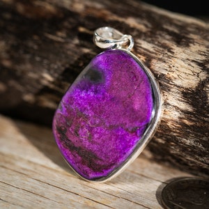 May include: A pendant with a vibrant purple gemstone, set in a silver bezel. The stone has dark, swirling patterns and a polished finish. The pendant is displayed on a wooden surface, showcasing its color and texture.