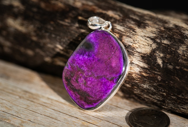 May include: A pendant with a vibrant purple gemstone, set in a silver bezel. The stone has dark, swirling patterns and a polished finish. The pendant is displayed on a wooden surface, showcasing its color and texture.