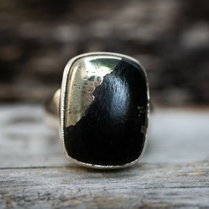 May include: A silver ring featuring a large, rectangular black stone with a metallic, reflective surface. The stone is set in a polished silver band, creating a striking contrast. The ring's design is simple yet bold.