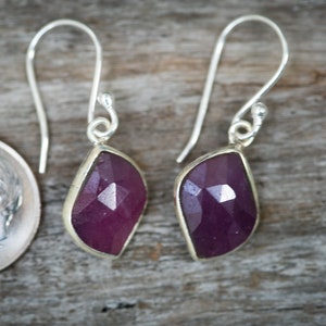 May include: A pair of silver dangle earrings with faceted purple gemstones. The earrings have a simple design and are perfect for everyday wear.