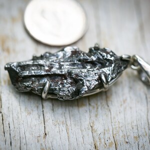 May include: A textured, irregularly shaped silver pendant with a loop for a chain. The pendant has a dark, metallic appearance with visible surface details. A silver coin is in the background.