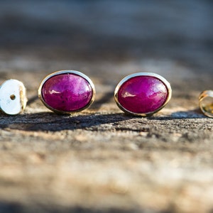 May include: A pair of oval-shaped ruby earrings with gold settings. The earrings are on a wooden surface.