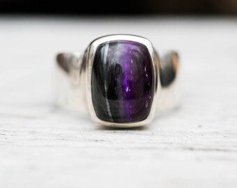 May include: A close-up of a silver ring with a rectangular, polished stone. The stone features a striking blend of purple, black, and gray hues. The ring's band is smooth and wide, creating a bold, modern aesthetic.
