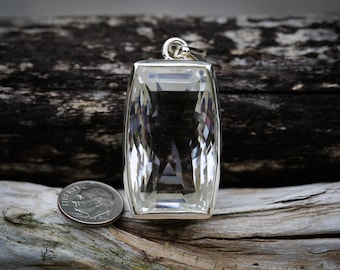 May include: A clear quartz crystal pendant with a silver bail. The pendant is rectangular in shape and has a faceted design. The pendant is sitting on a piece of driftwood next to a US dime for scale.