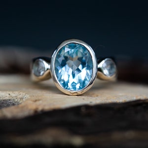 May include: A silver ring featuring a large oval blue topaz gemstone, flanked by two smaller round blue topaz gemstones.