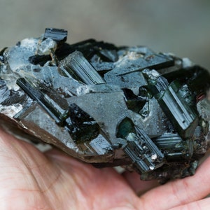May include: A dark green tourmaline crystal cluster with a rough, textured surface. The crystal formations are elongated and angular, with a glossy, reflective appearance. The mineral specimen is a deep, rich green colour.