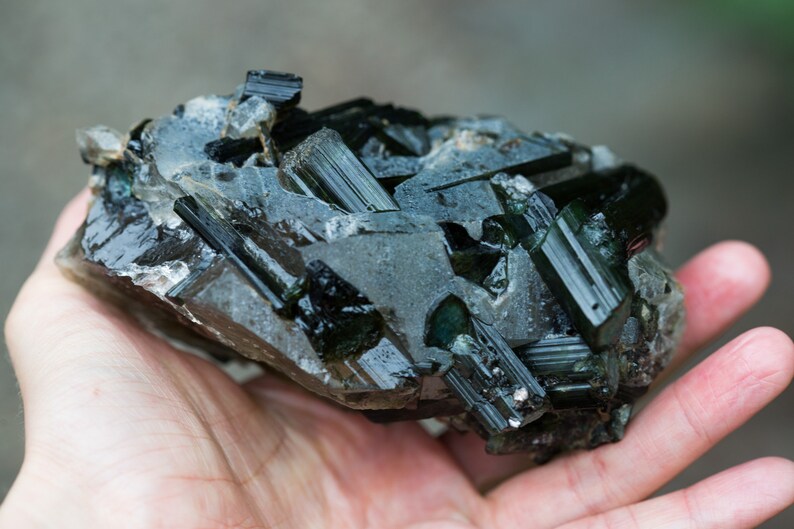 May include: A dark green tourmaline crystal cluster with a rough, textured surface. The crystal formations are elongated and angular, with a glossy, reflective appearance. The mineral specimen is a deep, rich green colour.