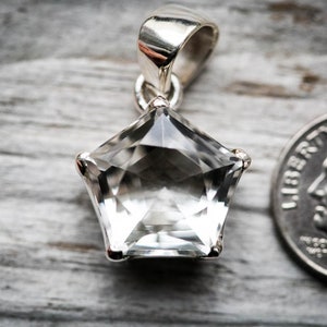 May include: A clear, faceted, star-shaped gemstone pendant set in a silver-toned metal frame with a bail at the top. A United States dime is positioned to the right of the pendant to provide a visual scale reference for the size of the item.