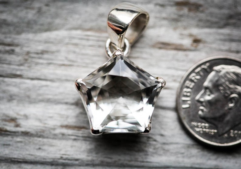 May include: A clear, faceted, star-shaped gemstone pendant set in a silver-toned metal frame with a bail at the top. A United States dime is positioned to the right of the pendant to provide a visual scale reference for the size of the item.