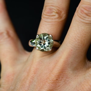 May include: A silver ring with a large, round, pale green gemstone. The ring is on a finger against a black backdrop. The gemstone is multi-faceted, reflecting light. The ring is a piece of jewellery.