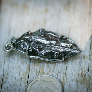May include: A silver pendant featuring a textured, irregularly shaped, dark gray stone. The stone is held in place by a silver wire frame. The pendant is displayed on a weathered wooden surface, with a coin in the foreground.