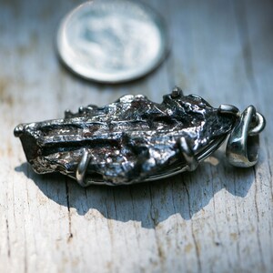 May include: A silver pendant with a textured, dark gray, irregular shape, possibly a meteorite. The pendant has a silver loop for a chain. A silver coin is in the background.