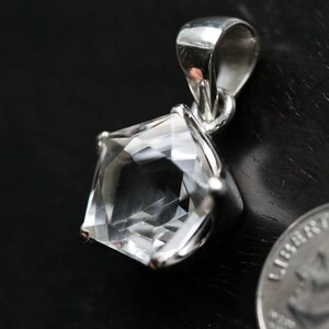 May include: A clear, faceted pentagonal gemstone pendant set in a polished silver-toned metal prong setting with a matching bail. A coin is positioned next to the pendant to provide a clear visual reference for its scale.