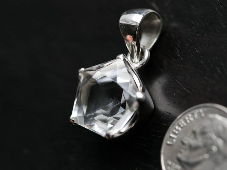 May include: A clear, faceted pentagonal gemstone pendant set in a polished silver-toned metal prong setting with a matching bail. A coin is positioned next to the pendant to provide a clear visual reference for its scale.