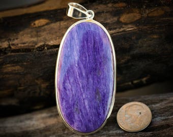 May include: A silver pendant featuring a large oval-shaped purple and blue gemstone with a swirling pattern. The pendant is on a wooden surface.