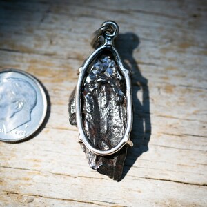 May include: A silver pendant with a dark, textured stone, possibly a meteorite, is displayed next to a U.S. dime. The pendant has a unique, irregular shape and is set in a silver frame with a loop for a chain. The background is a wooden surface.