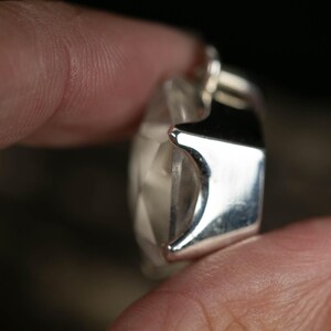 May include: A faceted clear gemstone pendant set in a polished silver metal frame. The stone features a geometric cut with multiple triangular surfaces that reflect light, held within a smooth, reflective silver setting with visible prongs.