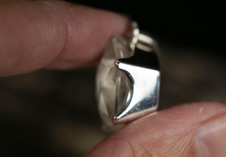 May include: A faceted clear gemstone pendant set in a polished silver metal frame. The stone features a geometric cut with multiple triangular surfaces that reflect light, held within a smooth, reflective silver setting with visible prongs.