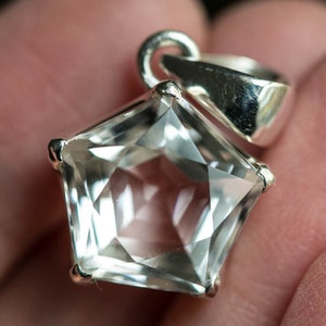 May include: A pentagonal clear gemstone pendant set in a polished silver metal frame. The faceted stone features a five-sided geometric shape with sharp edges and a reflective surface. A silver bail is attached to the top for hanging on a chain.
