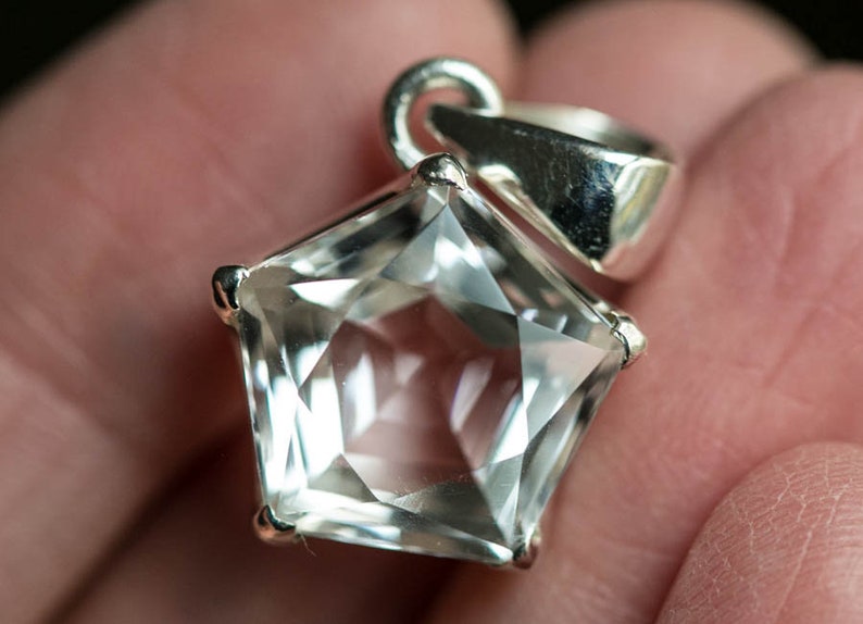May include: A pentagonal clear gemstone pendant set in a polished silver metal frame. The faceted stone features a five-sided geometric shape with sharp edges and a reflective surface. A silver bail is attached to the top for hanging on a chain.