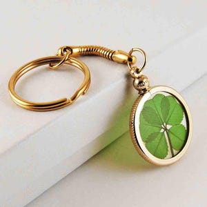May include: A gold-tone keychain with a four-leaf clover pressed inside a round glass locket. The keychain has a gold-tone split ring and a gold-tone chain.