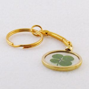 May include: A gold-toned keychain featuring a circular pendant with a ribbed edge. The pendant contains a preserved green four-leaf clover encased behind a clear surface. A flexible, coiled gold-toned metal connector links the pendant to a ring.