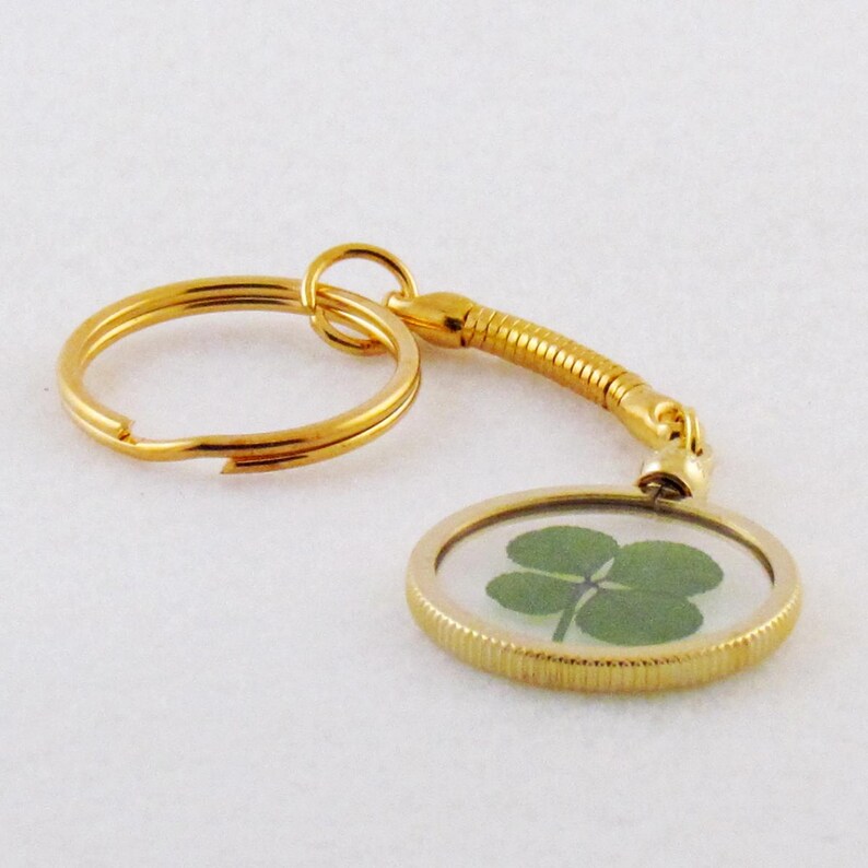 May include: A gold-toned keychain featuring a circular pendant with a ribbed edge. The pendant contains a preserved green four-leaf clover encased behind a clear surface. A flexible, coiled gold-toned metal connector links the pendant to a ring.