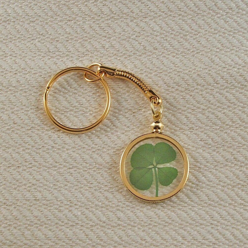 May include: A gold-toned metal keychain featuring a circular pendant with a clear center. Inside the pendant is a single pressed green four-leaf clover. The keychain includes a standard round split ring connected by a short, flexible metal coil.
