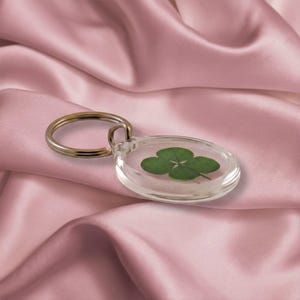 May include: A clear, oval-shaped keychain with a four-leaf clover encased inside. The keychain has a silver-colored metal ring. The background is a pink satin fabric.