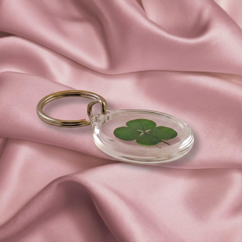 May include: A clear, oval-shaped keychain with a four-leaf clover encased inside. The keychain has a silver-colored metal ring. The background is a pink satin fabric.