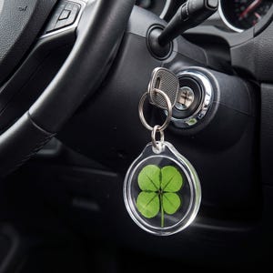 May include: A circular clear acrylic keychain containing a preserved green four-leaf clover. The keychain is attached to a silver metal split ring, which is connected to a silver car key inserted into the ignition of a black vehicle dashboard.