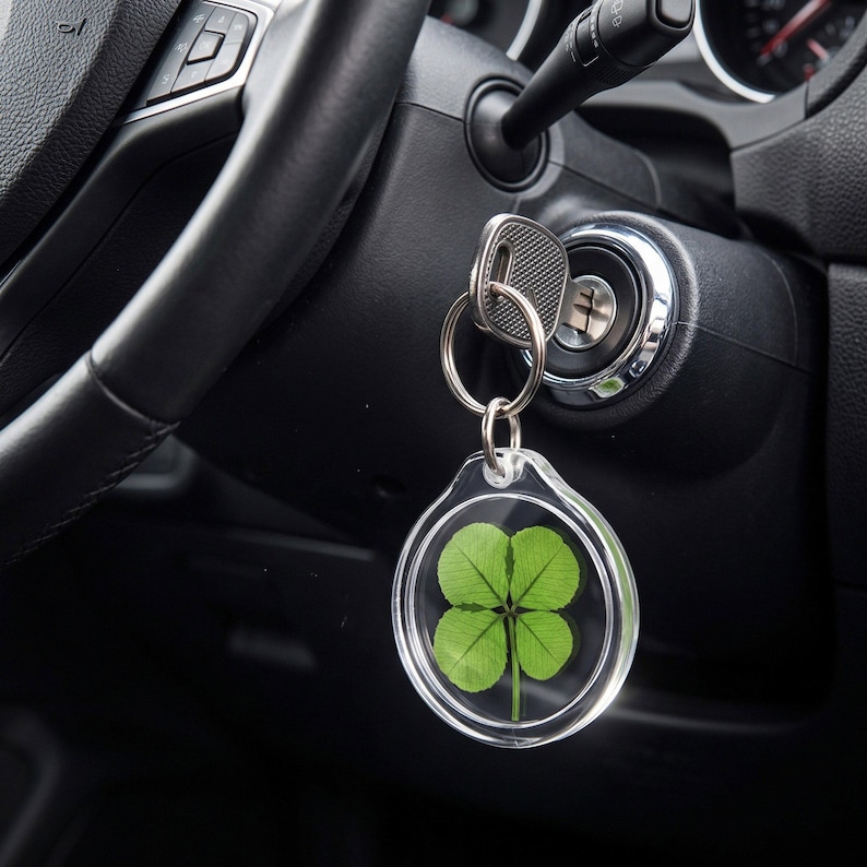 May include: A circular clear acrylic keychain containing a preserved green four-leaf clover. The keychain is attached to a silver metal split ring, which is connected to a silver car key inserted into the ignition of a black vehicle dashboard.