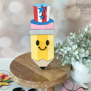 May include: A wooden pencil-shaped decoration with a smiling face, topped with a red, white, and blue Uncle Sam hat. The pencil is yellow with a gray band and black tip. The hat has blue stars. The words "browncow creatives handmade" are visible.