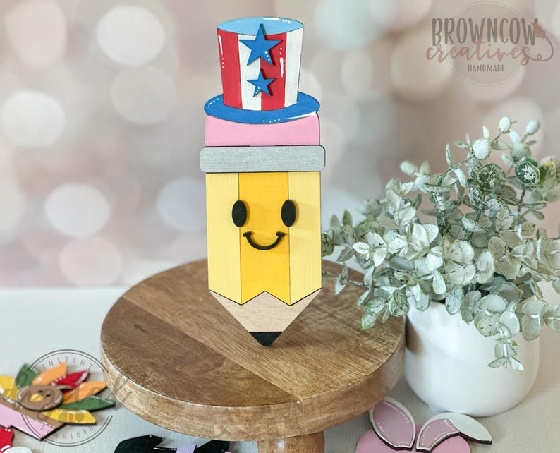May include: A wooden pencil-shaped decoration with a smiling face, topped with a red, white, and blue Uncle Sam hat. The pencil is yellow with a gray band and black tip. The hat has blue stars. The words "browncow creatives handmade" are visible.
